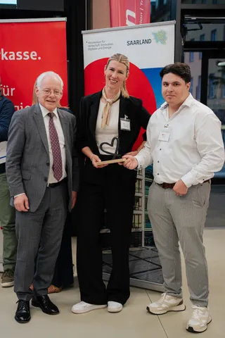 Tarik Ermis met twee andere personen op het podium van Demo Day 2026, met de hartvormige trofee van de publieksprijs